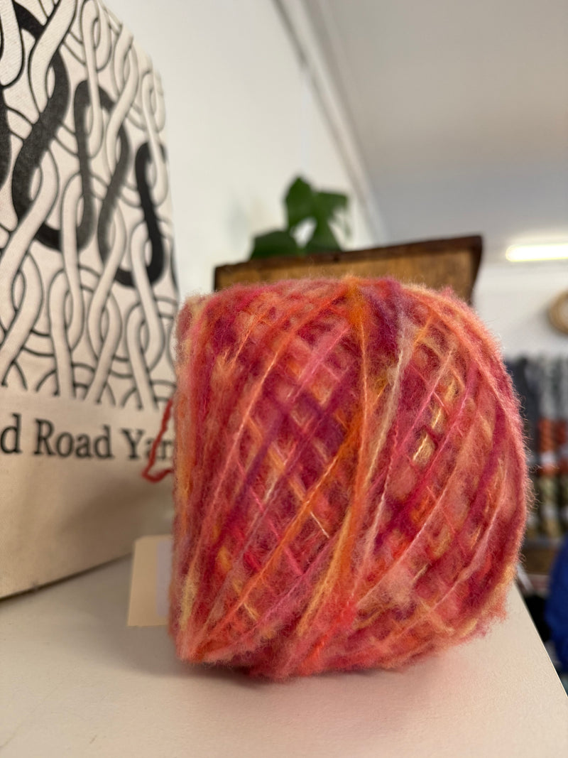 A ball of variegated merino boucle yarn in shades of yellow, orange, and red, displayed on a white surface next to a canvas tote bag with black knit stitch patterns and the text 'Holland Road Yarn'.