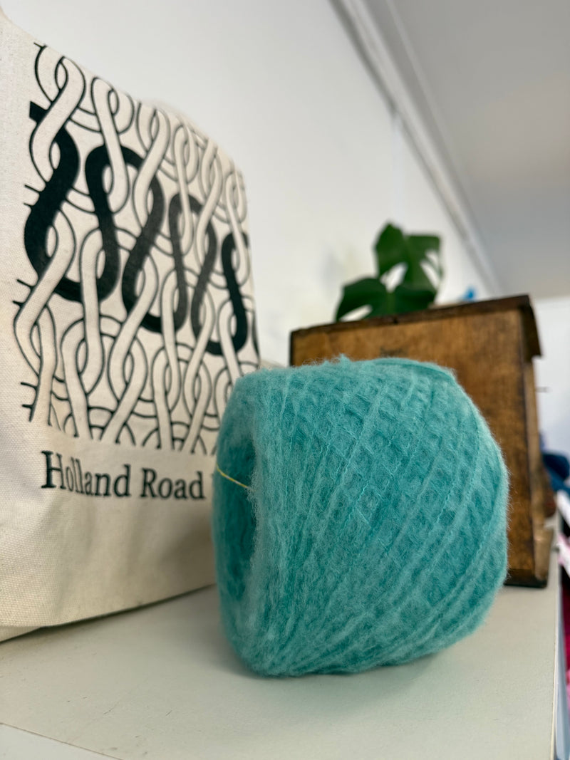 A ball of fluffy teal blue yarn sits on a white surface in a styled indoor setting, with a canvas tote bag and a wooden box blurred in the background.