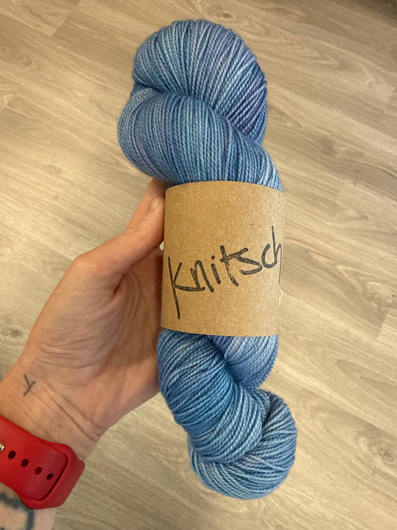 A skein of tonal blue-purple high twist merino sock yarn, with Knitsch branding. 