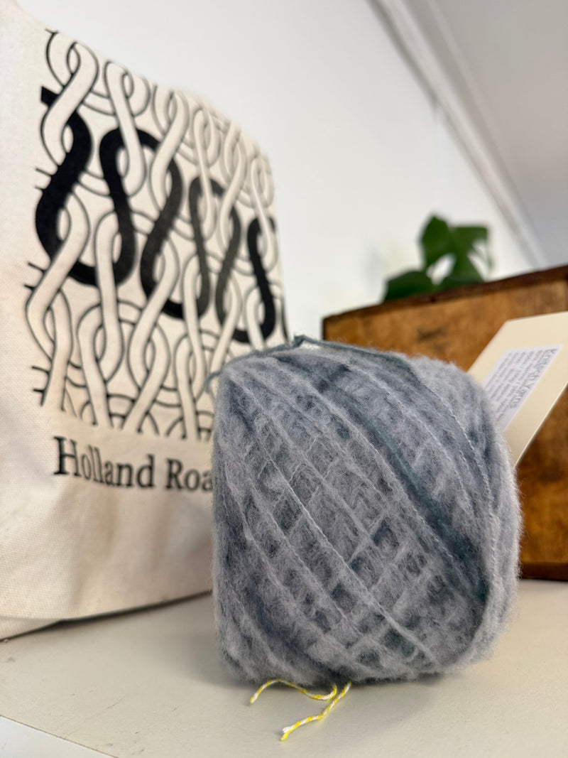 A ball of fluffy gray yarn sits on a tabletop in front of a cream tote bag with black Celtic knot artwork and the text 'Holland Road'. merino boucle. A wooden box and blurred greenery are in the background.