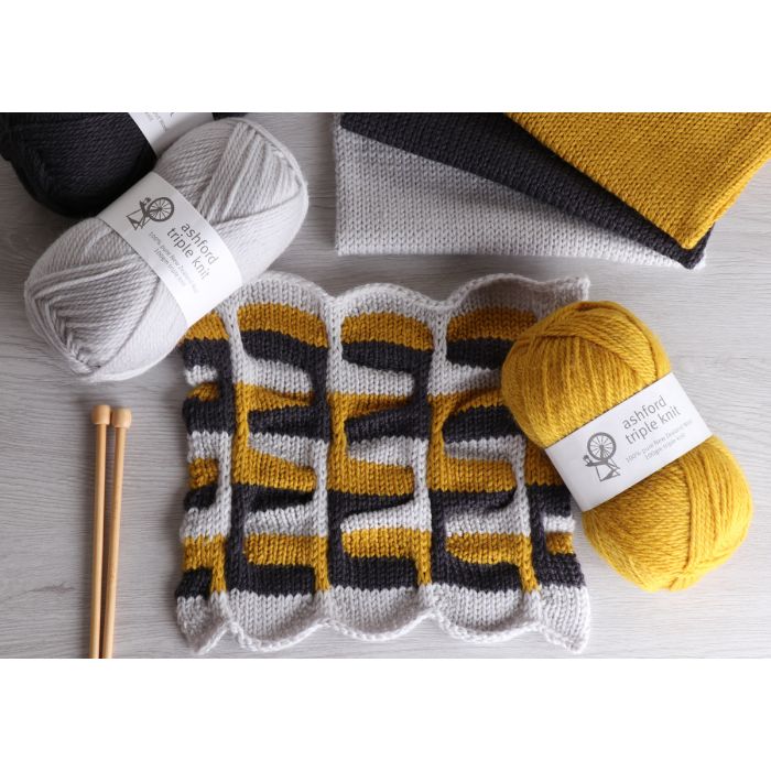 A photo showing a partially knitted fabric with a pattern, alongside two skeins of yarn in white and yellow colors, and a knitting needle.