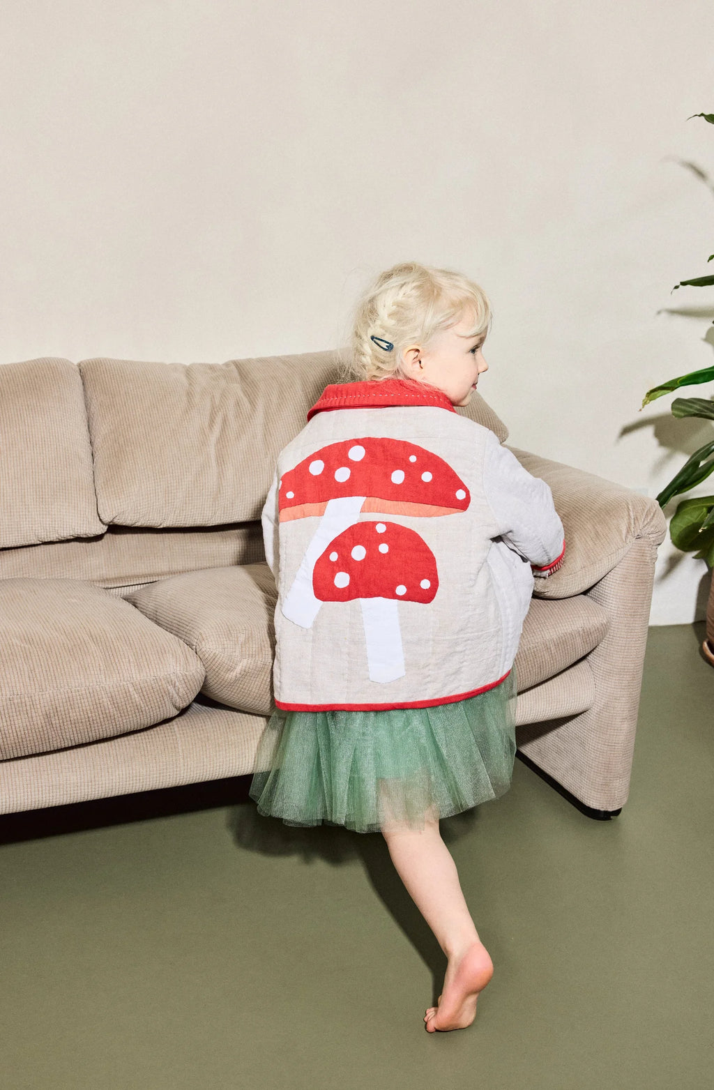 Child wearing a mushroom-patterned jacket sitting on a couch in a living room.