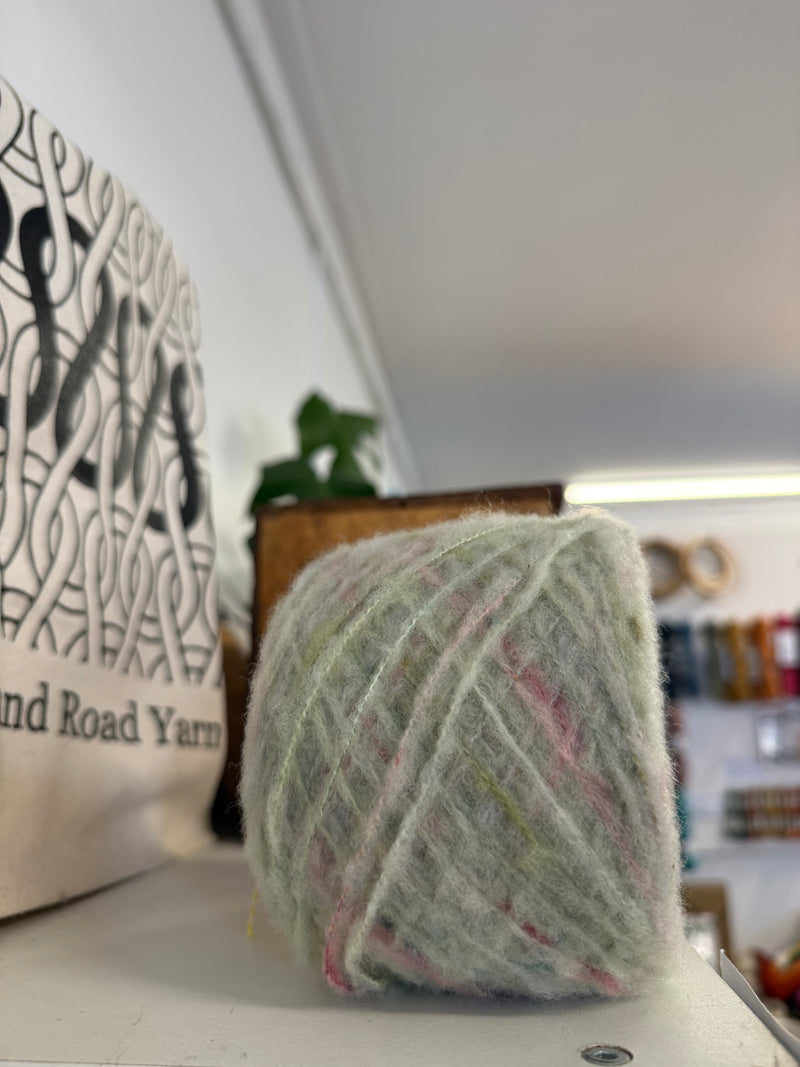 A ball of fluffy, variegated yarn in pale sage green, pink, and cream sits on a white surface in a cozy, softly blurred craft room setting. A canvas bag with the text 'Holland Road Yarn' is visible on the left.