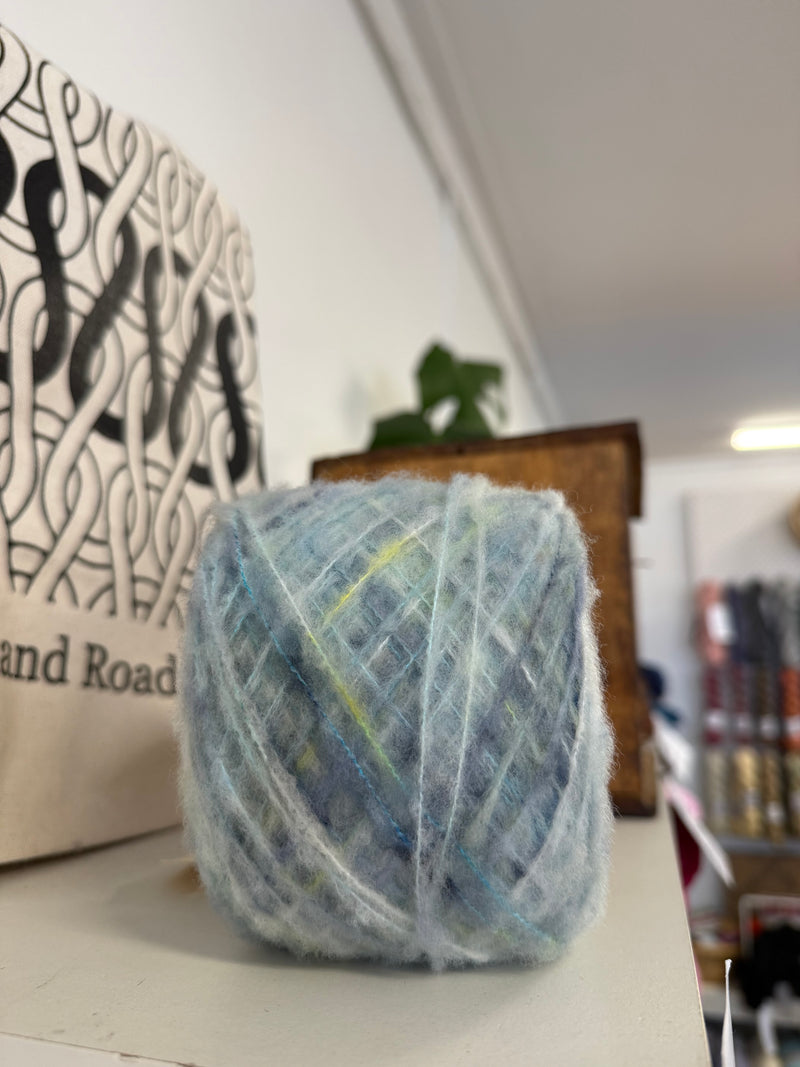 A ball of variegated merino boucle yarn in shades of light blue, white, and soft green yellow, sitting on a shelf next to a canvas tote bag.