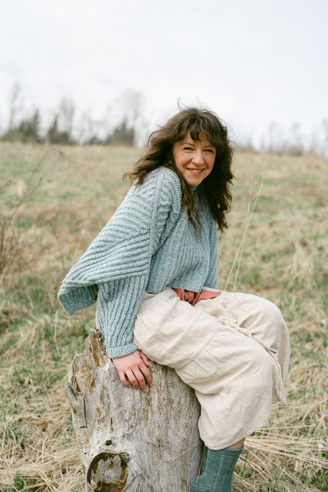 Woman sitting on a log in a field wearing a blue sweater and beige pants.