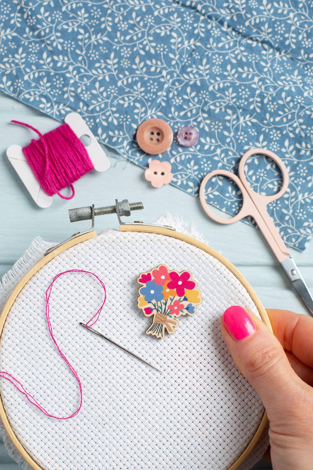Embroidery hoop with floral needle minder, pink thread, scissors, and buttons on a blue floral fabric background.