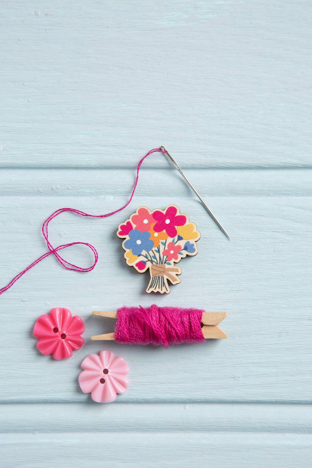 Decorative needle minder with flowers, pink thread, and a needle on a light blue wooden surface.