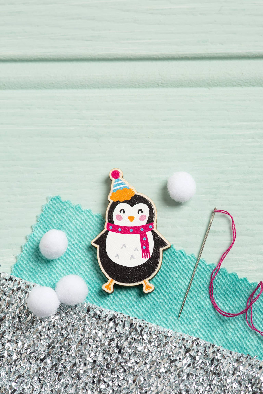 Decorative penguin needle minder with a party hat on a textured surface with sewing materials.