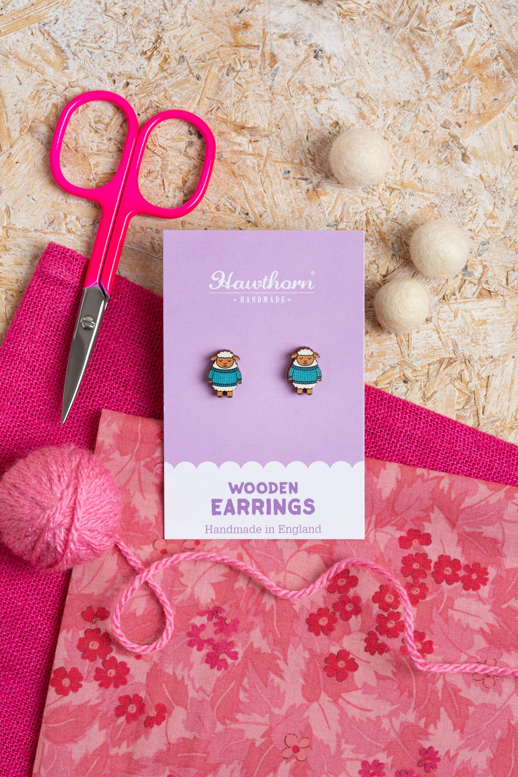 Wooden earrings on a card with pink yarn and scissors on a textured surface