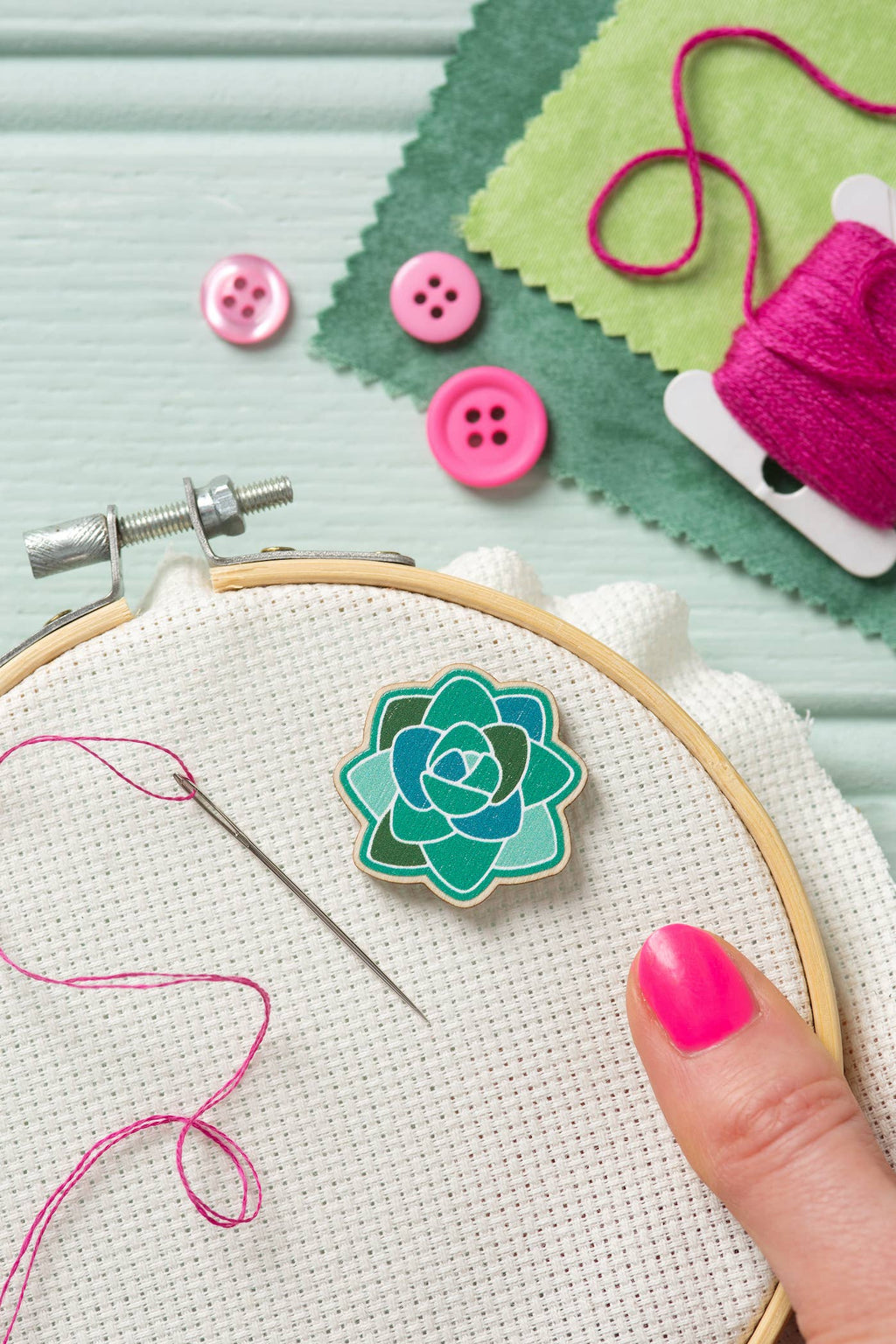 Embroidery hoop with a green succulent needle minder, pink buttons, and thread on a light blue background.