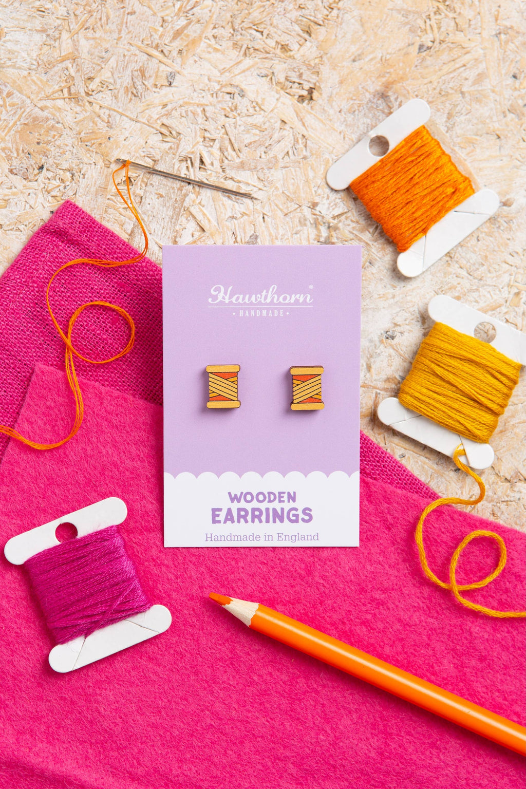 Wooden earrings on a card with spools of thread and a pencil on a wooden surface