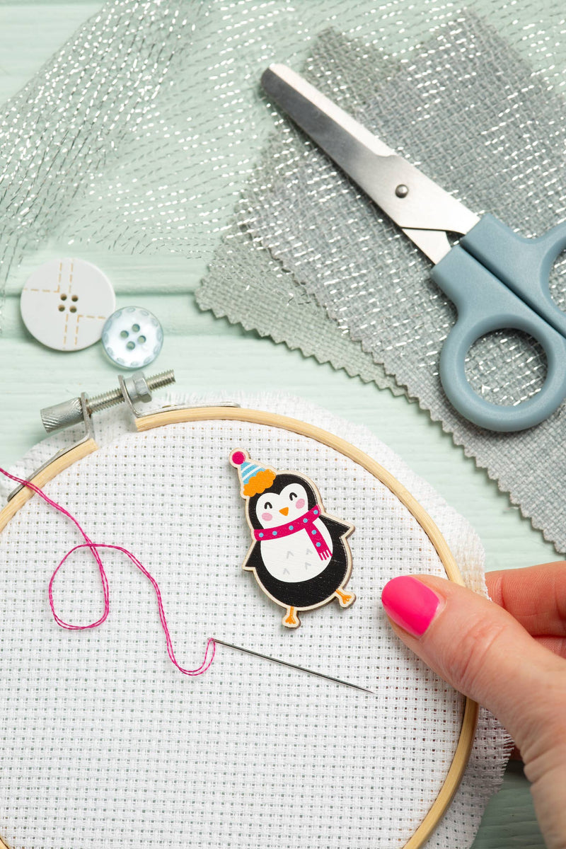 Cross-stitching project with a penguin needle minder, scissors, and embroidery hoop on a light green surface.