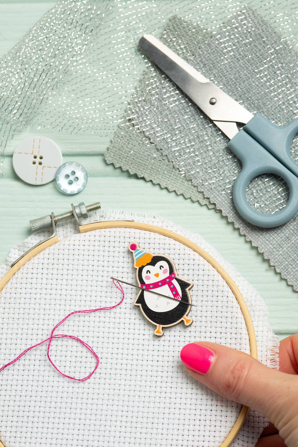 Embroidery hoop with a penguin needle minder, scissors, and thread on a light green background
