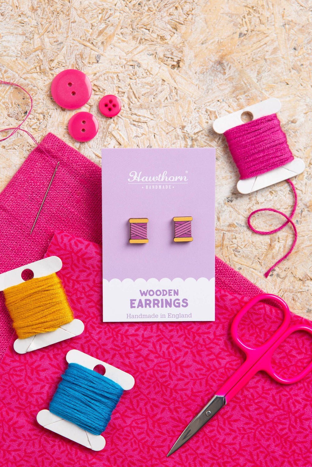Handmade wooden earrings packaging with pink thread spools, scissors, and buttons on a textured surface.