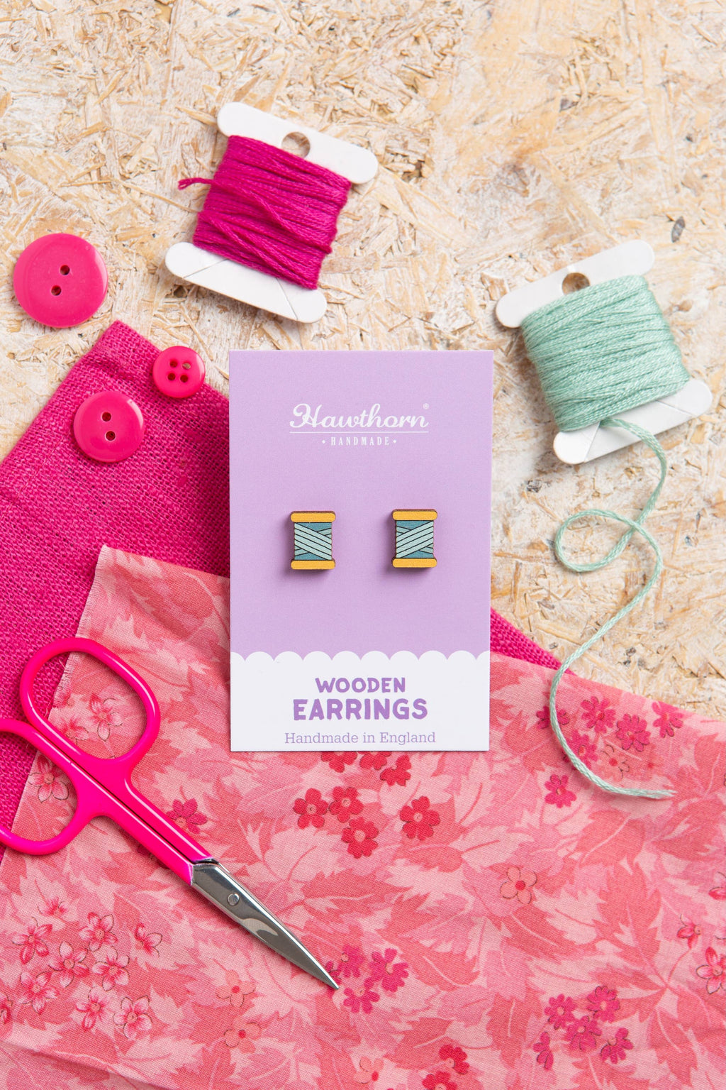 Wooden earrings on a card with pink fabric, scissors, and spools of thread on a wooden surface.