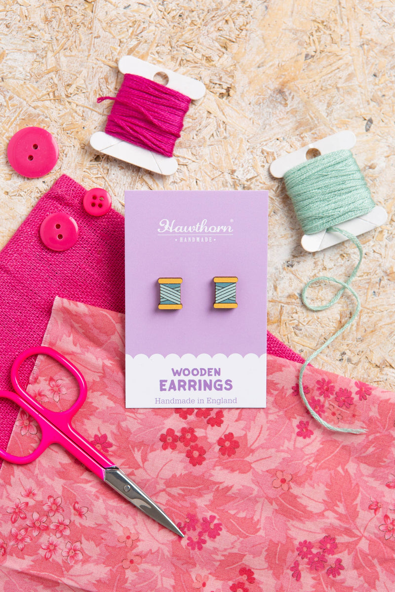 Wooden earrings on a card with pink fabric, scissors, and spools of thread on a wooden surface.