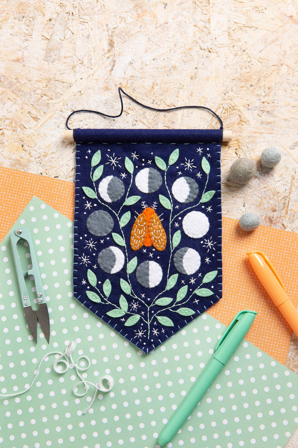Decorative felt banner with moon phases and a butterfly on a textured surface with crafting tools.