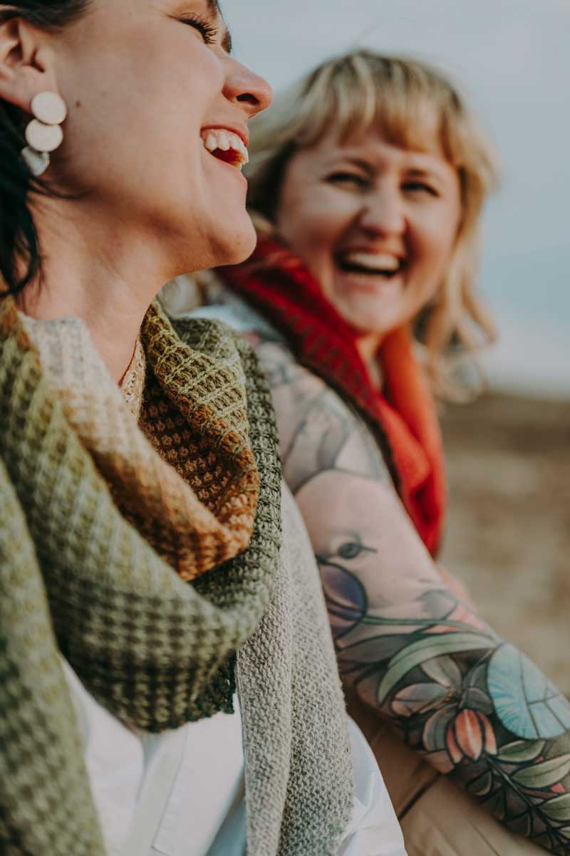 Two women laughing outdoors, one with a green shawl and visible tattoos.