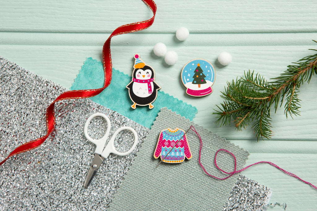 Decorative pins with penguin and sweater design, scissors, and festive elements on a light blue background.