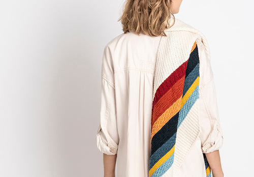 Person wearing a white blouse with a colorful striped hand knit shawl on the back against a plain background