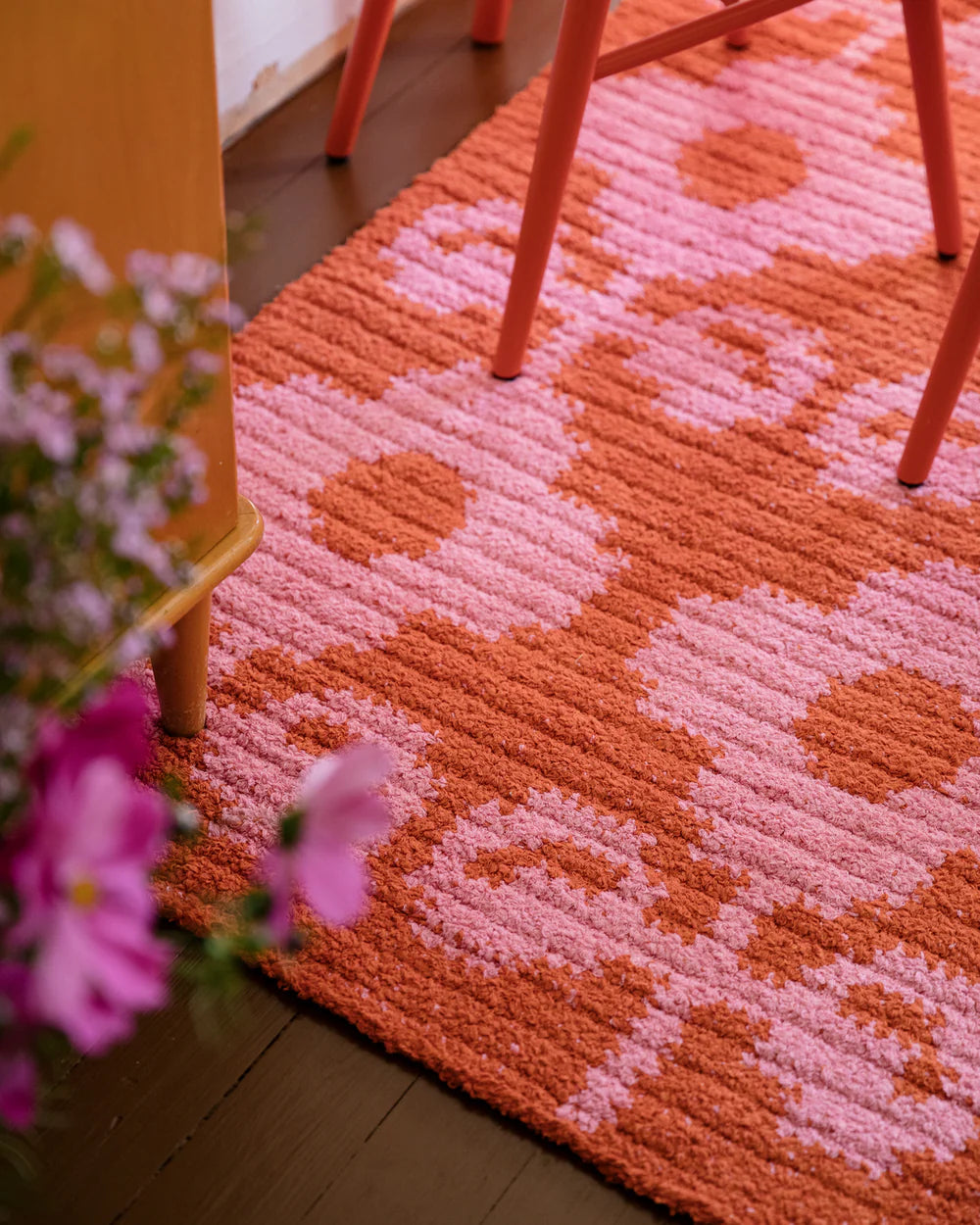 Pink and orange patterned crochet rug with a chair and flowers in the foreground